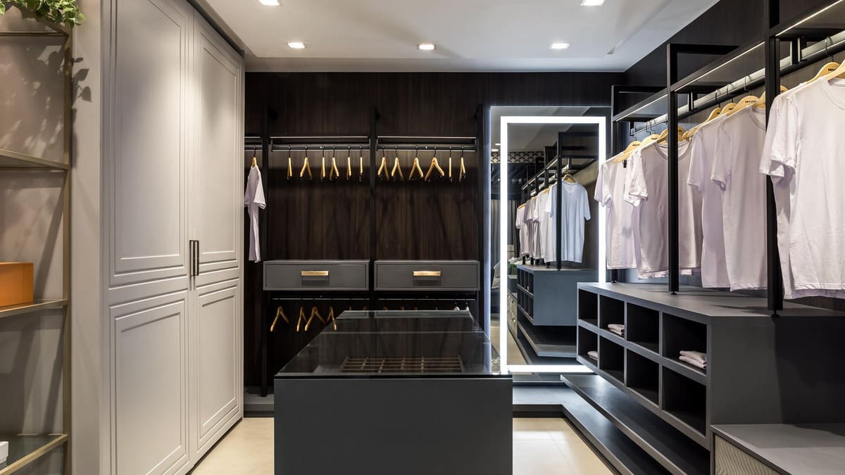 Walk-in closet with dark open wardrobes, a glass-top island and a lit mirror