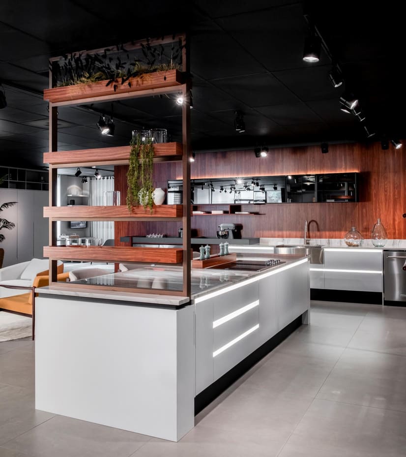 White kitchen island with lit drawer pulls under a wood and metal shelving unit with plants
