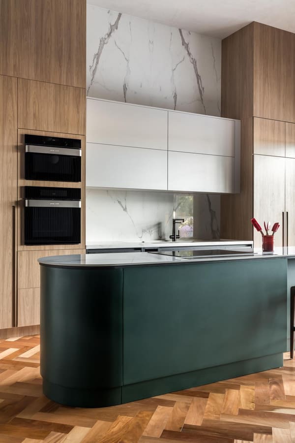 Curved green kitchen island on a herringbone wood floor, with marble-backed tall cabinets